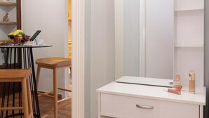 A room with a white vanity, a mirror, a small table with a bowl of fruit, and a chair. - Acropolis Promenade by Teknys Hosting (Athina)