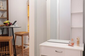 A room with a white vanity, a mirror, a small table with a bowl of fruit, and a chair. - Acropolis Promenade by Teknys Hosting (Athina)