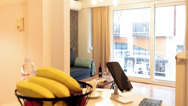A room with a desk, a computer, a bowl of fruit, a bottle, a wine glass, and a sofa. - Acropolis Promenade by Teknys Hosting (Athina)