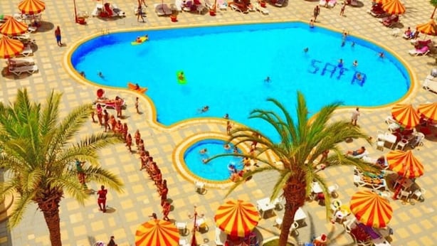 A swimming pool with sun loungers and umbrellas, surrounded by palm trees and a tiled deck.