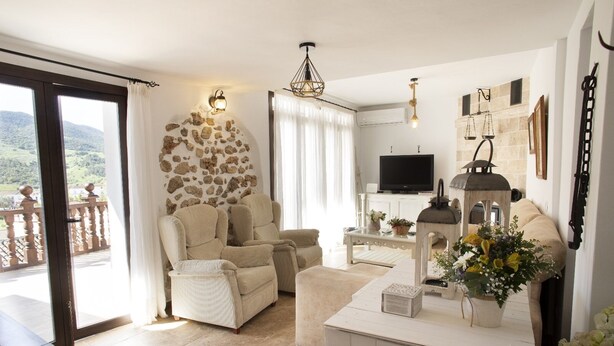 A living room with a stone wall, a television, a fireplace, and a balcony with a view of the hills.