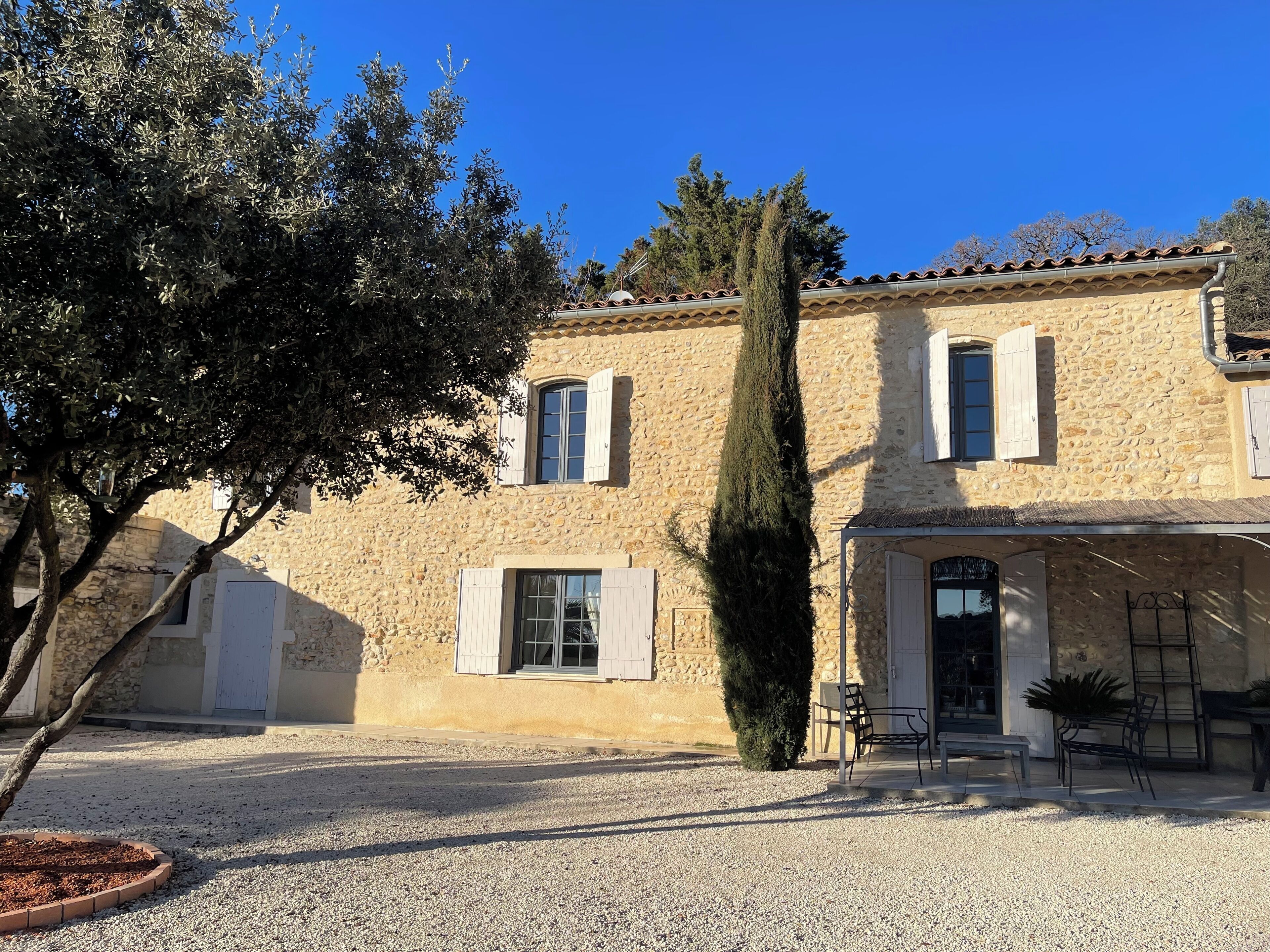 Un bâtiment en pierre avec une allée gravillonnée, un grand arbre et des sièges à l’extérieur.