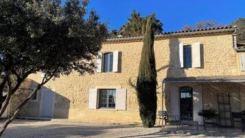 Un bâtiment en pierre avec une allée gravillonnée, un grand arbre et des sièges à l’extérieur.