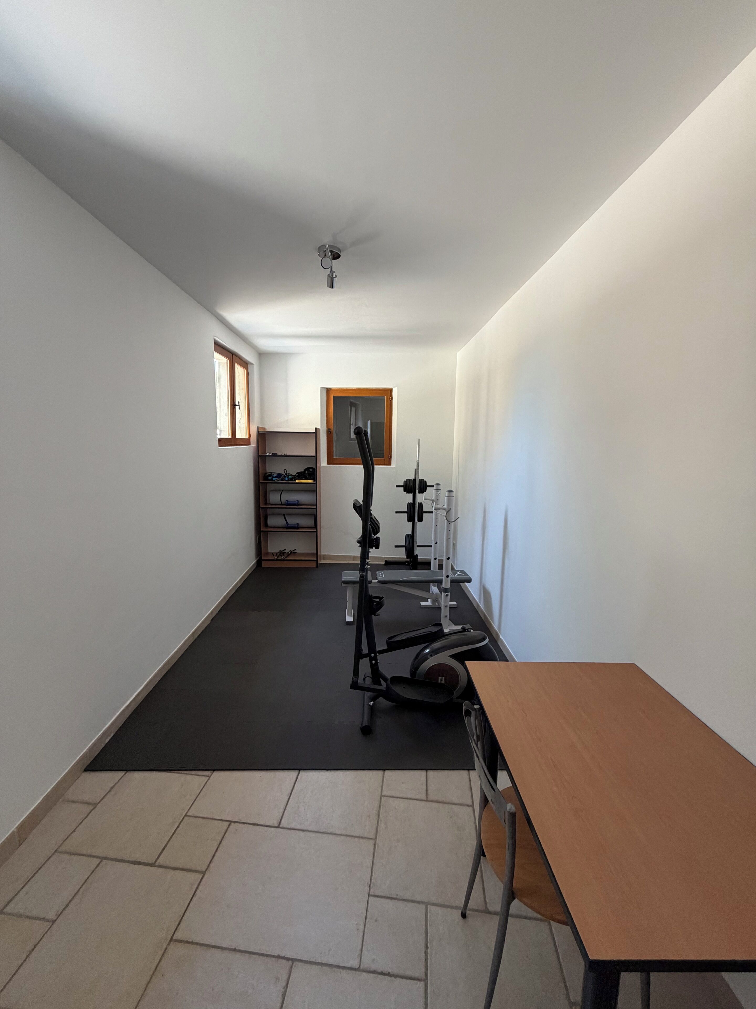 Ein Zimmer mit Fitnessgeräten, einem Holztisch und einem Fenster.