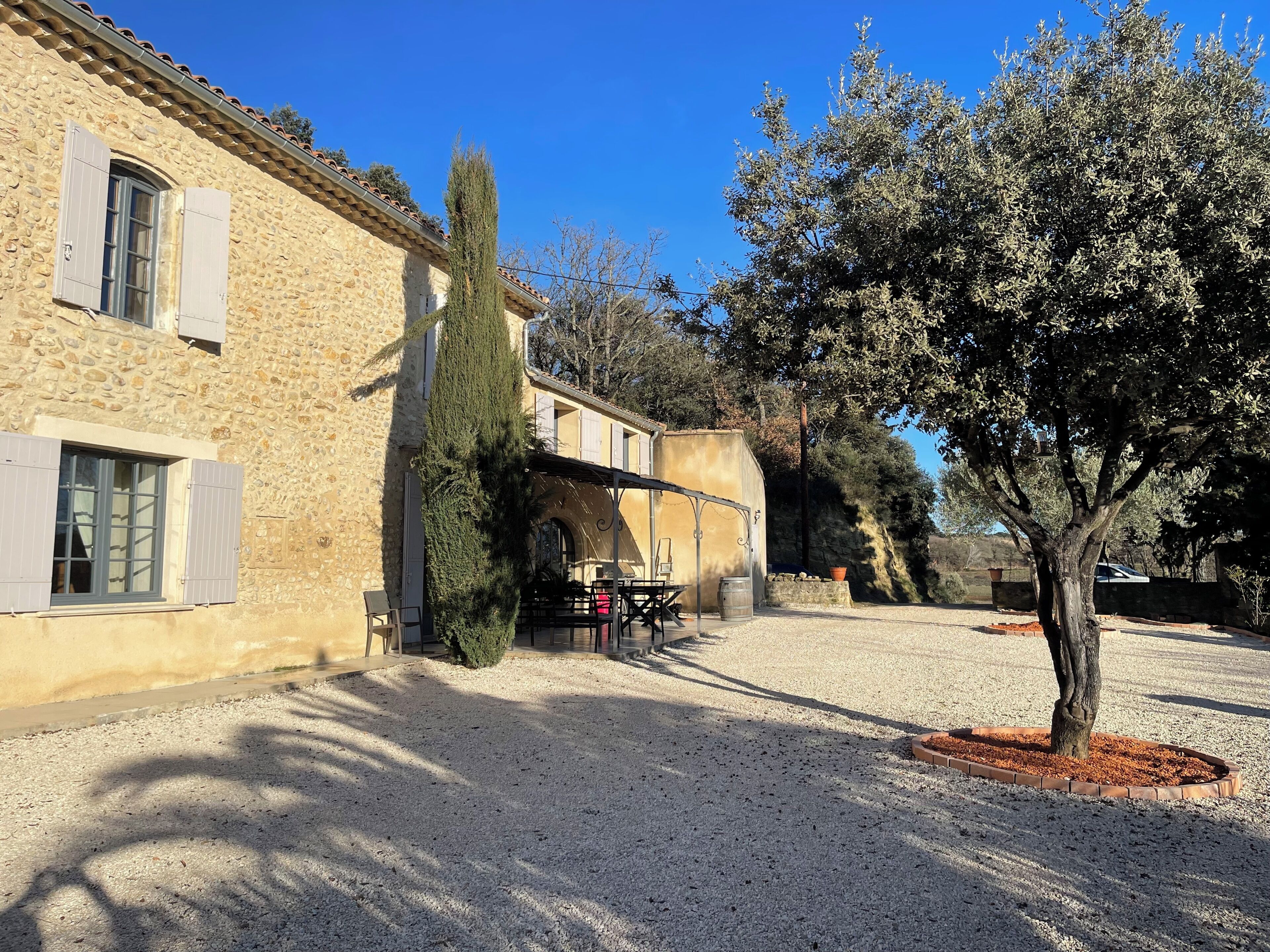 Un bâtiment en pierre avec une cour recouverte de gravier, un grand arbre et une tonnelle en métal.
