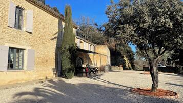 Un bâtiment en pierre avec une cour recouverte de gravier, un grand arbre et une tonnelle en métal.