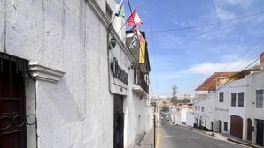 A narrow street with white buildings, a visible storefront with a sign, and a clear sky. - Hostal Orosheam (Arequipa)