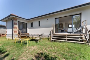 Una casa moderna con un amplio ventanal corredero, un patio con mobiliario exterior y un interior visible que incluye una cama y un comedor.
