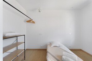 A small, sparsely furnished room with a bunk bed, a wooden shelf, a hanging light fixture, and a single wall socket.