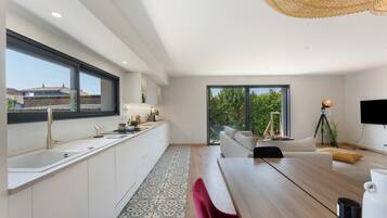 Een moderne keuken met een groot raam, een eethoek met een houten tafel, en een zithoek met een bank en een televisie.