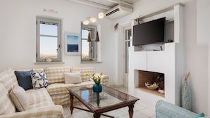 A living room with a checkered sofa, a glass coffee table, a television mounted above a fireplace, and a view of the sea through a window. - Villa Kohyli by Mediterà (Paros)