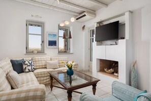 A living room with a checkered sofa, a glass coffee table, a television mounted above a fireplace, and a view of the sea through a window. - Villa Kohyli by Mediterà (Paros)