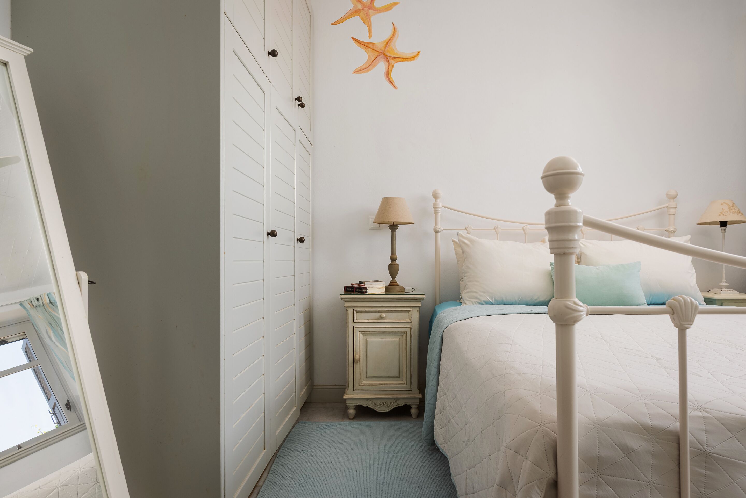 A bedroom with a white bed, a nightstand with a lamp, a dresser, and starfish wall art.