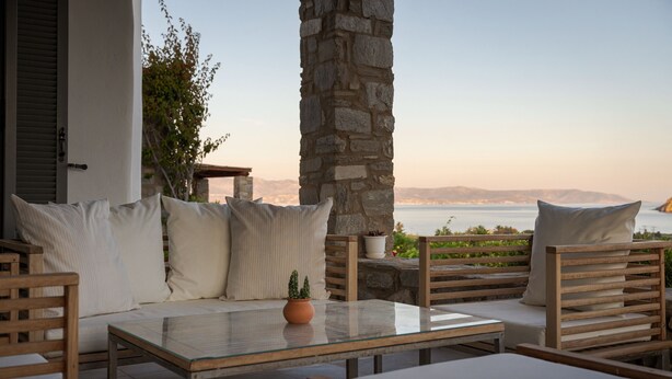 A sunlit outdoor seating area with a stone pillar, cushioned chairs, a glass-top table, and a view of the sea. - Villa Kohyli by Mediterà (Paros)