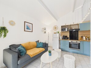 A modern apartment with a grey sofa, a small round table, a white coffee table, a kitchen with blue cabinets, and a white refrigerator.