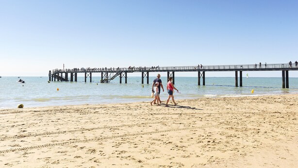 Una playa con un muelle que se adentra en el agua, gente caminando sobre la arena y barcos a lo lejos.