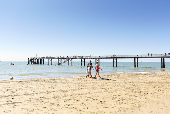 Une plage avec une jetée qui s’avance dans l’eau, des gens qui se promènent sur le sable et des bateaux à l’horizon.