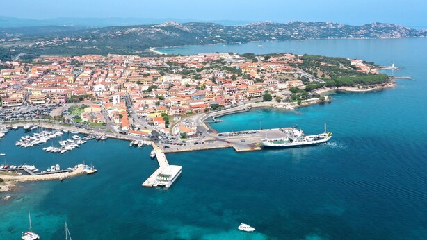 Vista aerea di una località costiera con un porto turistico, barche e un faro.