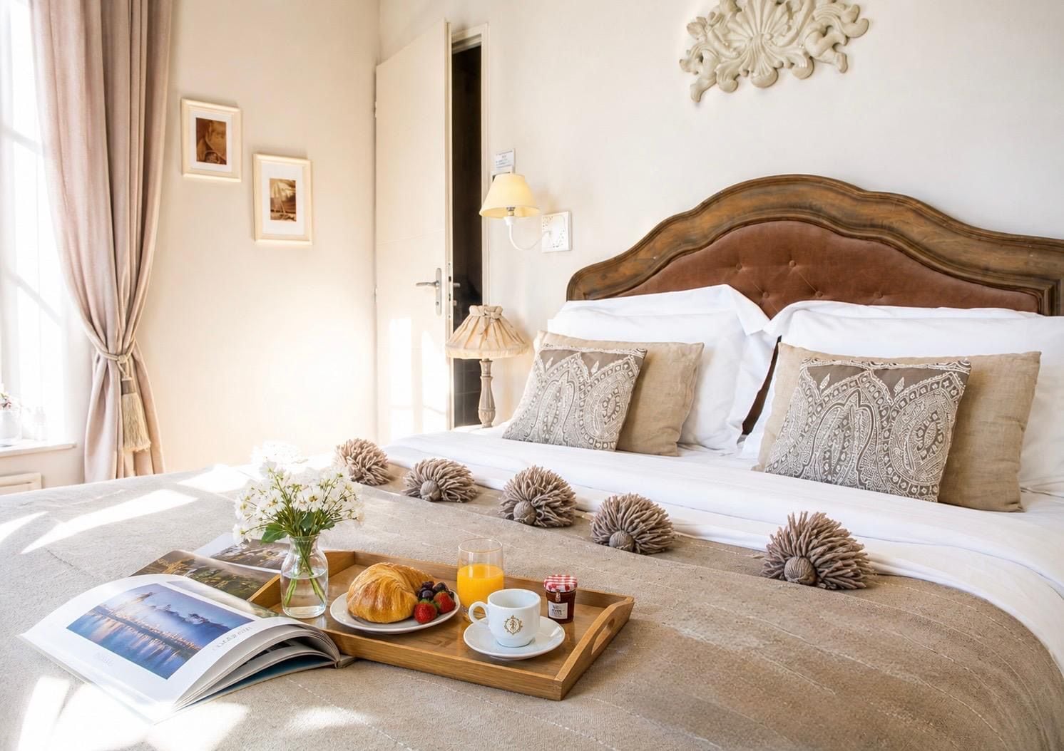 Cama bem arrumada com cabeceira de madeira, mesa de cabeceira com abajur e bandeja com itens de café da manhã.