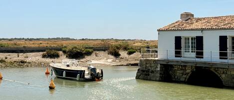 A boat docked near a white building with a tiled roof, a stone bridge, and a sandy shoreline.