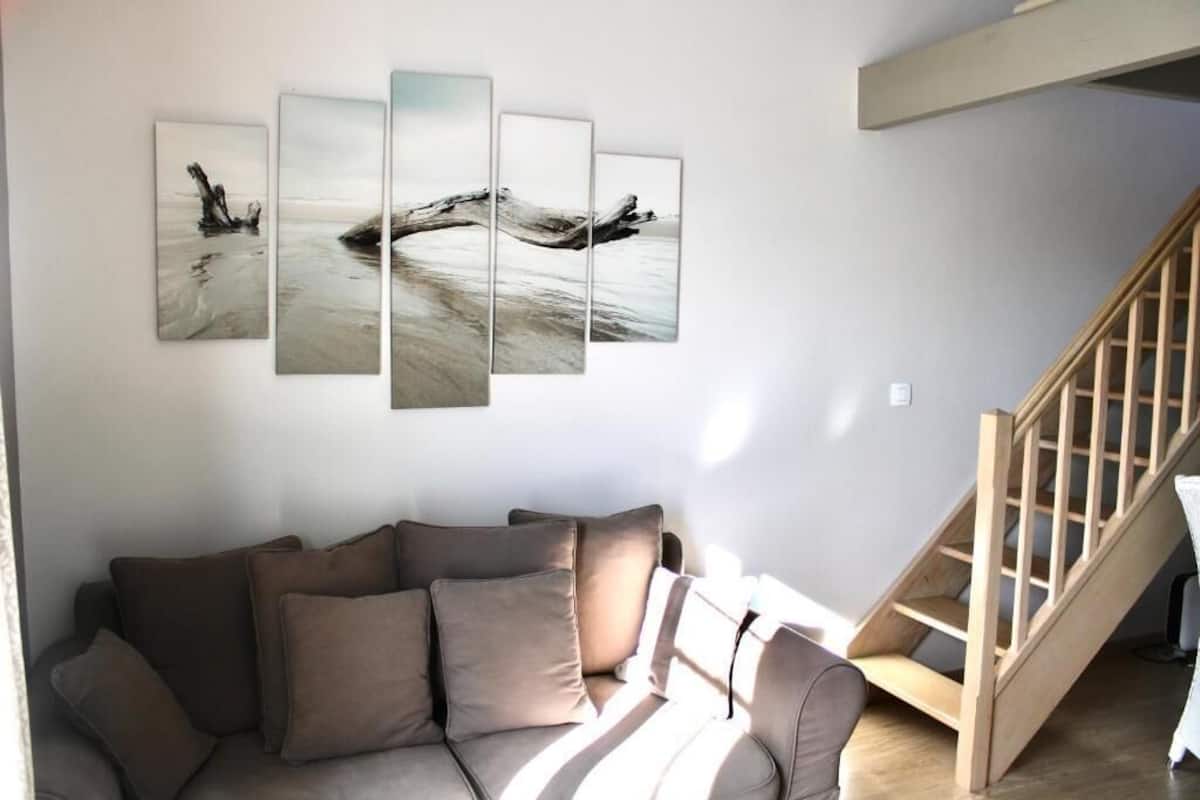 A living room with a sofa, a wooden staircase, and a wall with a multi-panel artwork.