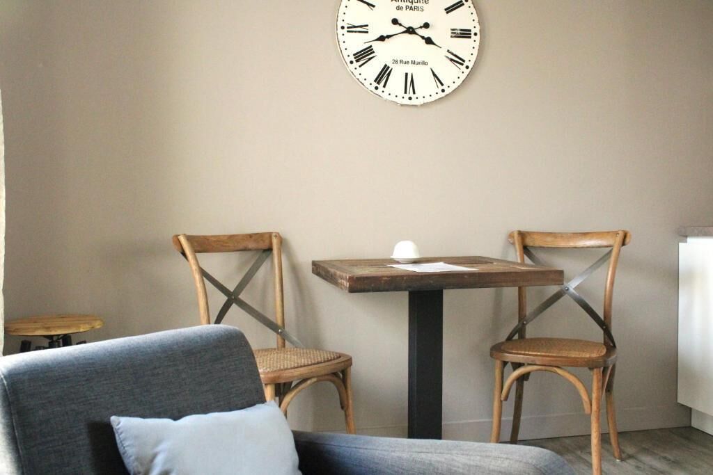 A room with a large wall clock, two wooden chairs, a small table, and a sofa.