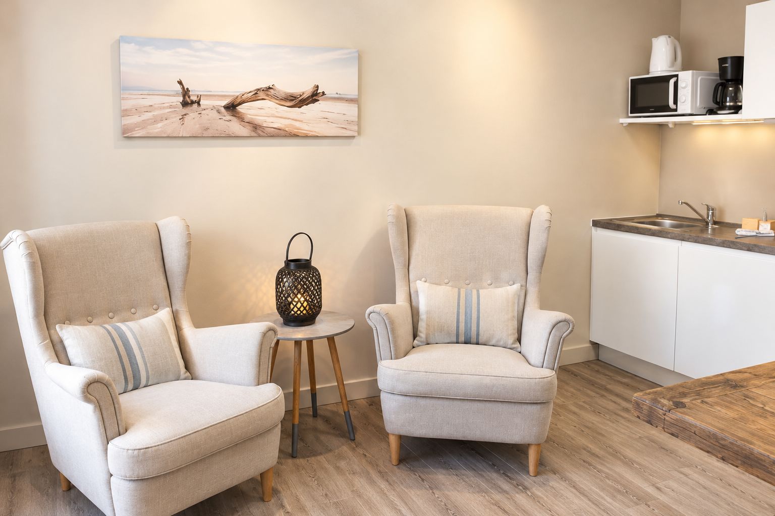 Une cuisine moderne équipée d’un four à micro-ondes et d’une machine à café, deux fauteuils avec des coussins à rayures, une petite table en bois surmontée d’une lanterne, et un tableau représentant un paysage désertique accroché au mur.
