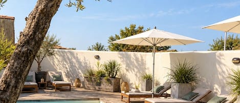 Zona de piscina con sillas de descanso y sombrillas, rodeada por un muro bajo y plantas en macetas.