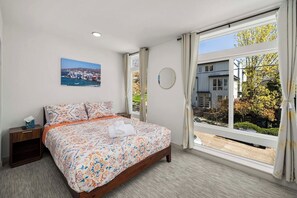 A bedroom with a large bed, a bedside table, a window with curtains, and a wall-mounted round mirror. - Brand New 2BR Townhome in Fremont | Rooftop Patio (Seattle)