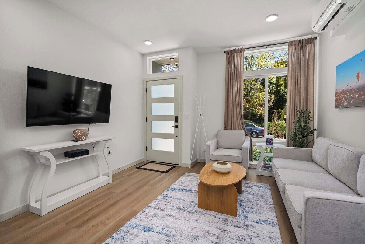 Una sala de estar moderna con un televisor de pantalla plana, una consola blanca, un sofá beige, una mesa de centro y un ventanal con cortinas.