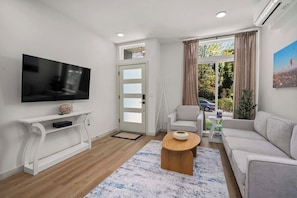 Ein modernes Wohnzimmer mit Flachbildfernseher, weißem Konsolentisch, beigefarbenem Sofa, Couchtisch und großem Fenster mit Vorhängen.