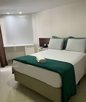 Een keurig opgemaakt bed met een groene sprei, een nachtkastje met een telefoon, en een raam met gordijnen.