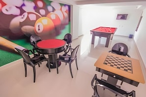 Een biljart, een pooltafel en een schaaktabel in een ruimte met een groen vloerkleed en witte muren.