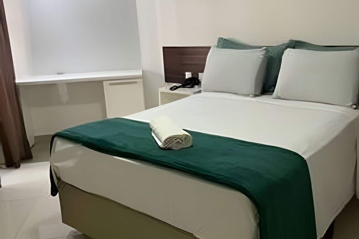 A neatly made bed with a green bedspread, a bedside table with a phone, and a window with curtains.