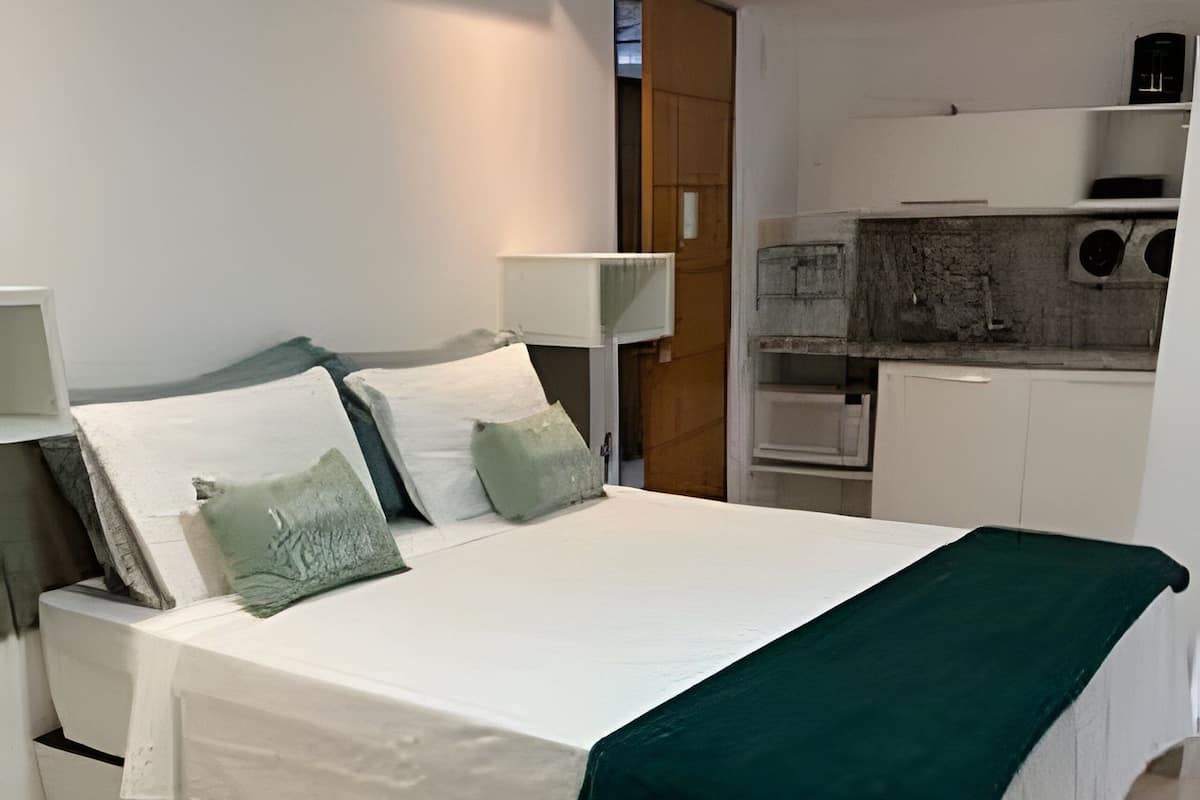 A neatly made bed with pillows and a green throw blanket, a white bedside table, a lamp, and a kitchenette in the background.