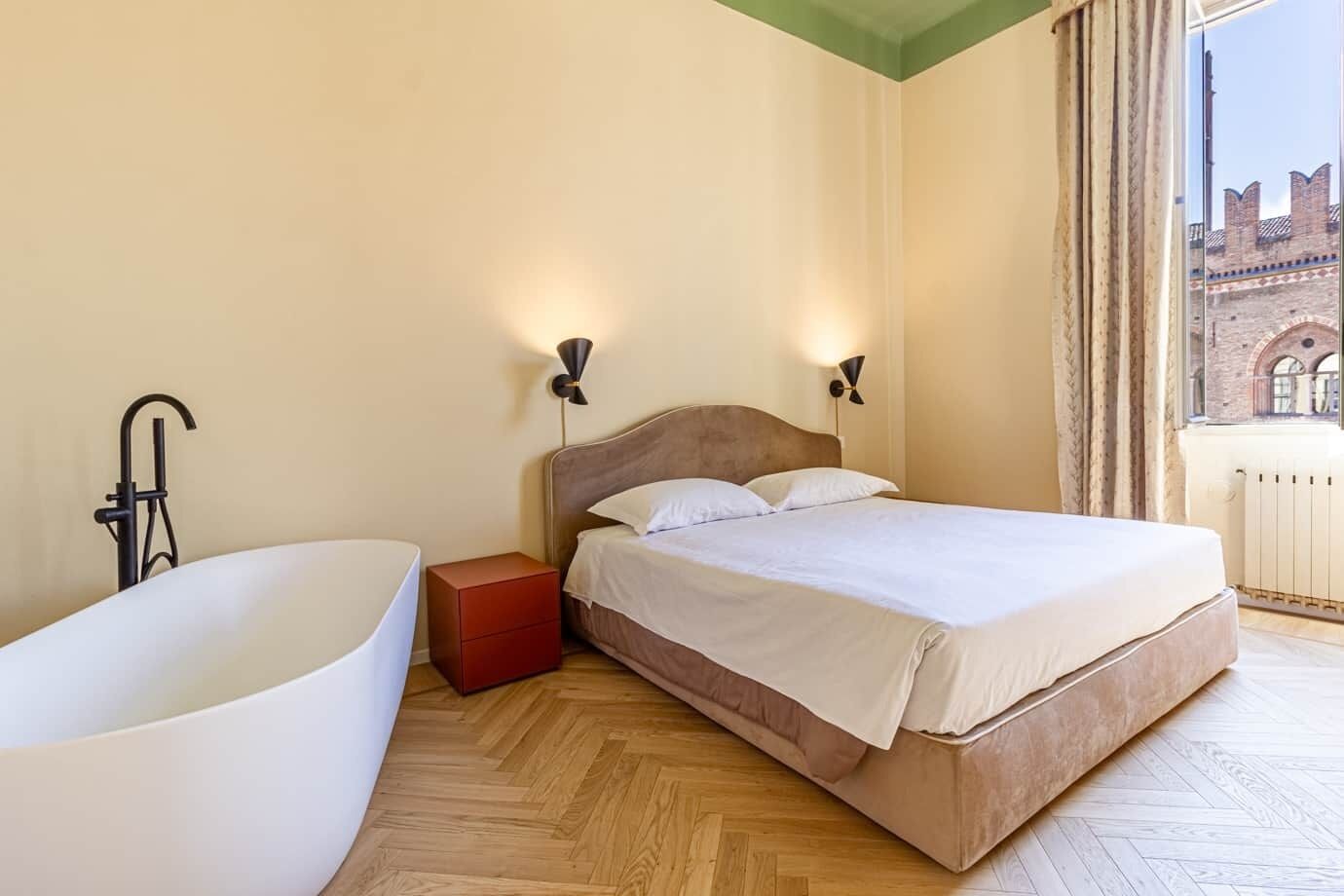 A bedroom with a large bathtub, a wooden floor, a bed with white linens, two wall-mounted lamps, and a view of a building through a window.