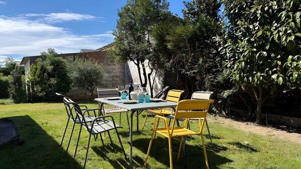 Un espace de restauration en plein air avec une table et des chaises, entouré de verdure et avec un bâtiment en arrière-plan.
