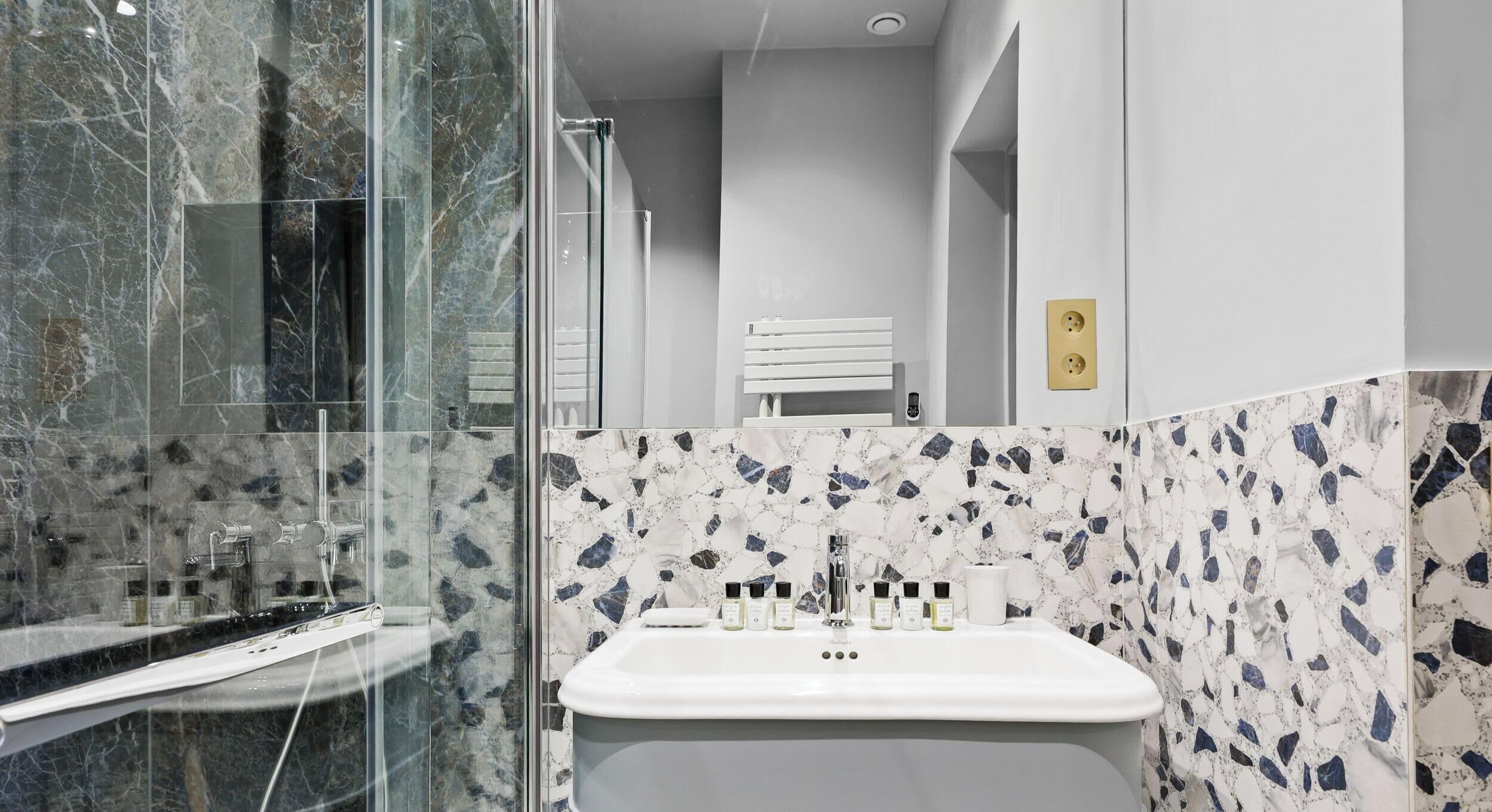 Une salle de bain dotée d’une douche à parois de verre, d’un lavabo blanc avec des articles de toilette et d’un pare-baignoire en marbre.