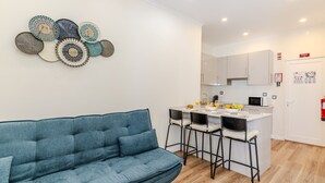 Een moderne woonkamer met een blauwe bank, een keuken met witte kasten, en een eethoek met barkrukken.