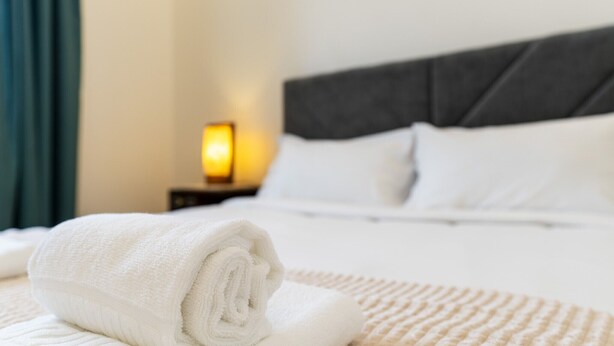 Een netjes opgemaakt bed met witte lakens, een opgerolde handdoek, een bedlampje en een donker hoofdeinde.