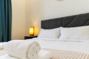 Una cama bien hecha con sábanas blancas y una toalla enrollada, una lámpara de noche y un cabecero oscuro.
