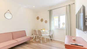 A living room with a sofa, a dining table with chairs, a TV mounted on the wall, and a window with curtains. - #379 Sunny Corner | Albufeira Stay (Albufeira)