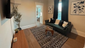 Ein Wohnzimmer mit einem dunklen Sofa, einem Couchtisch, einem an der Wand montierten Fernseher und einer Zimmerpflanze.