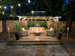 Un patio al aire libre con una pérgola, luces colgantes, un pozo de fuego y mobiliario exterior.