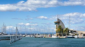 A marina with boats docked, a modern building with a curved roof, and palm trees. - La Frégate by Interhome (Port Camargue)