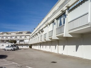 Ein modernes Gebäude mit weißer Fassade, mehreren Balkonen und einem Parkplatz mit einigen Autos.