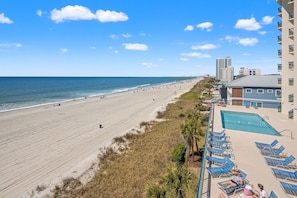 Blick auf einen Strand mit Poolbereich, Liegestühlen und einem mehrstöckigen Gebäude.