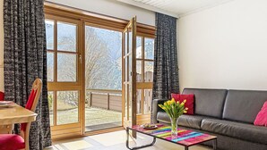 A living room with a sofa, a coffee table with a colorful top, a chair, and a view through a glass door to a snowy outdoor area. - Bernkogel by Interhome (Saalbach-Hinterglemm)