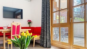 A modern living room with a red sofa, a wooden dining table, a TV mounted on the wall, and a large window with curtains. - Bernkogel by Interhome (Saalbach-Hinterglemm)