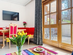 A modern living room with a red sofa, a wooden dining table, a TV mounted on the wall, and a large window with curtains. - Bernkogel by Interhome (Saalbach-Hinterglemm)